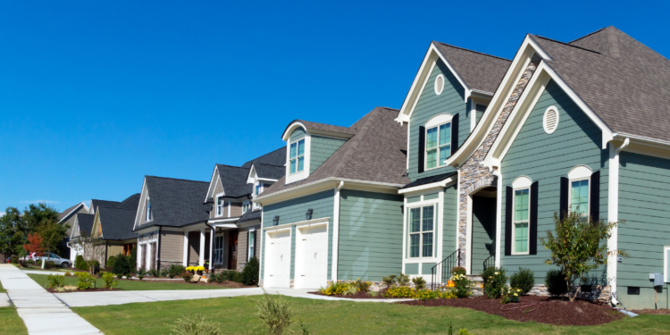A row of modern suburban houses in a uniform architectural style along a sidewalk and lawn under a clear blue sky