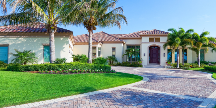 A modern Florida-style home with palm trees, lush landscaping, and a wide brick driveway under a clear blue sky